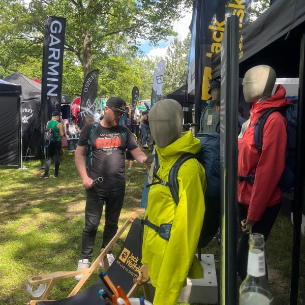 Mann in schwarzem Shirt steht vor einem Stand mit Outdoor-Bekleidung und Markenflaggen im Hintergrund