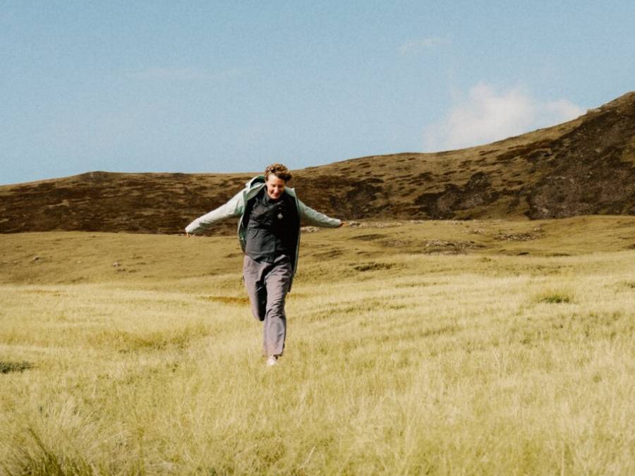 Mann in sommerlicher Wanderkleidung rennt mit ausgestreckten Armen über ein Feld